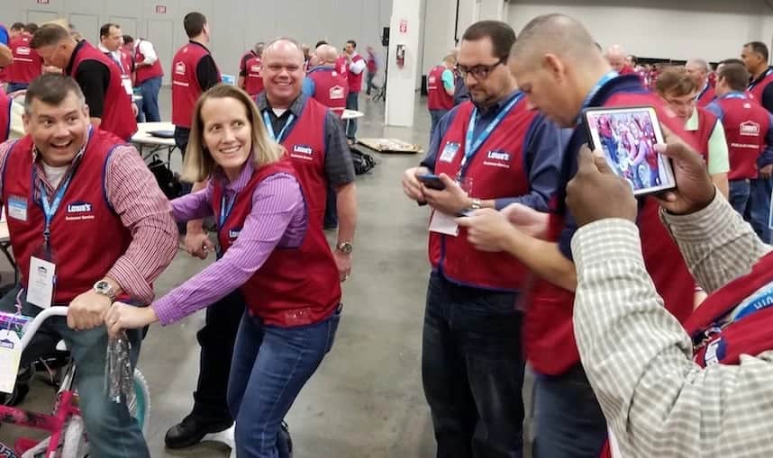 Lowe's managers in red vests on the Sands Expo floor during the Bike Build Donation, one sitting on a child's bike