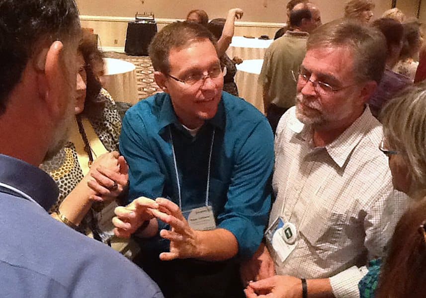 Four coworkers in a tight huddle mid-argument over a plan during Competition to Collaboration