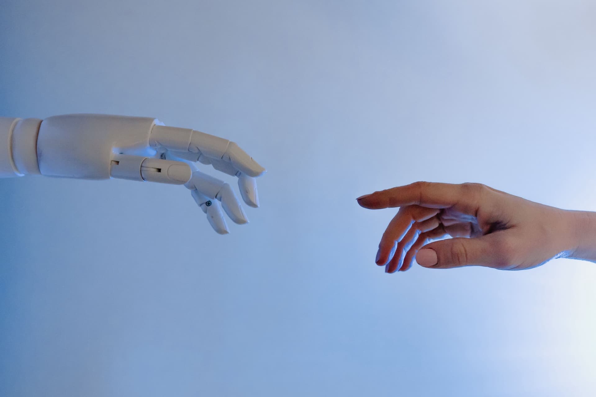 A white robotic hand and a human hand reaching toward each other against a pale blue background