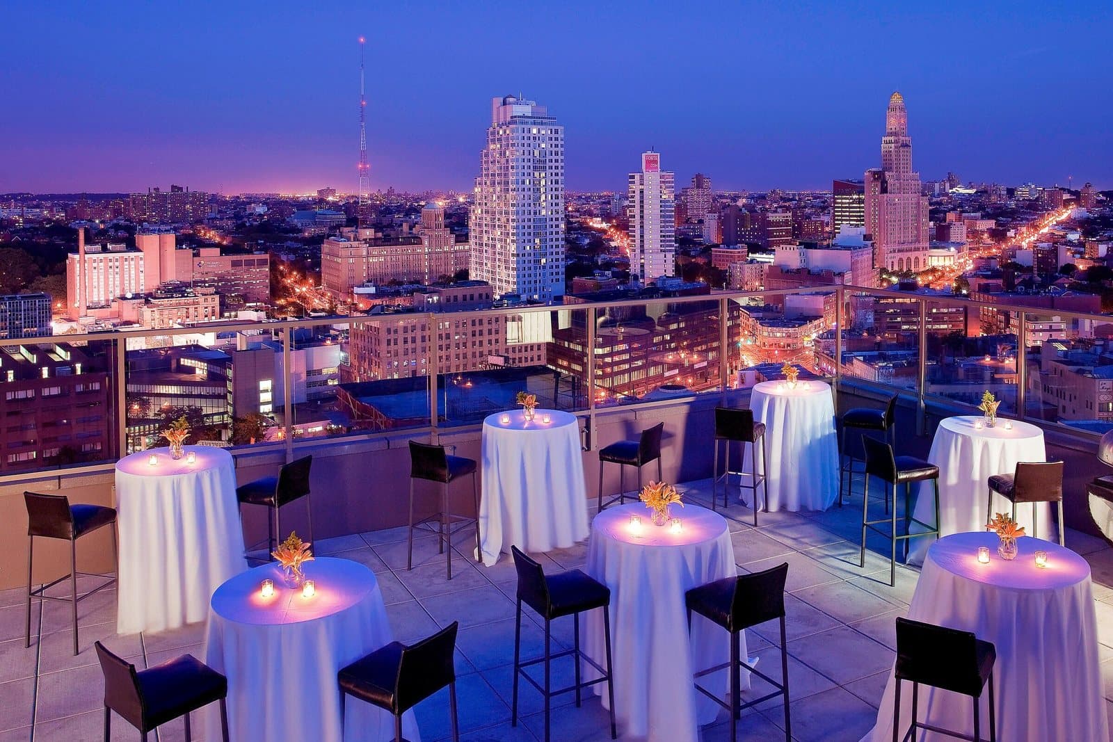 Cocktail tables set on the Sheraton Brooklyn New York's rooftop at dusk, with Downtown Brooklyn lit behind