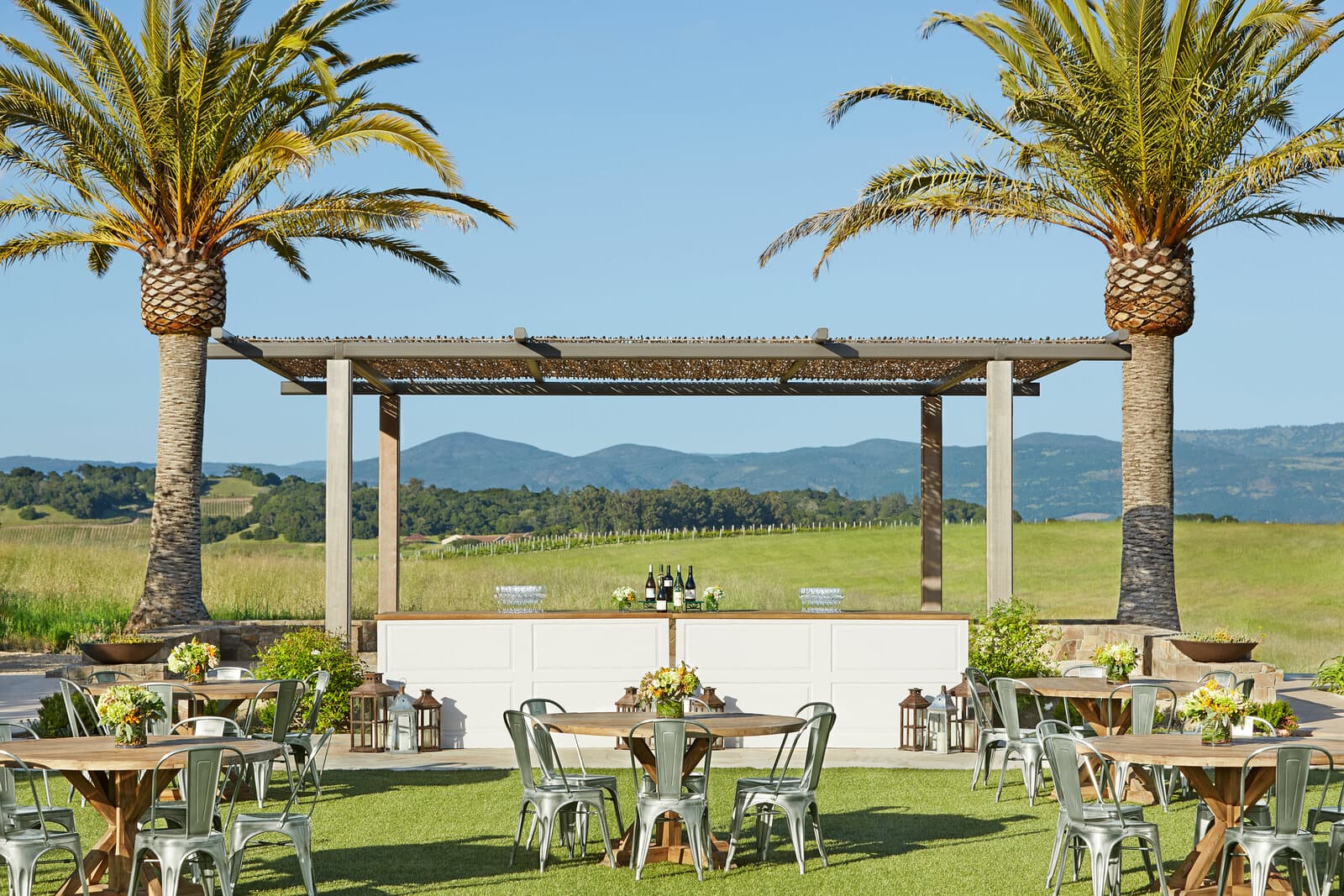 Farm tables and chairs on the lawn beneath a pergola framed by palm trees, with vineyard hills behind, at Carneros Resort and Spa in Napa, CA