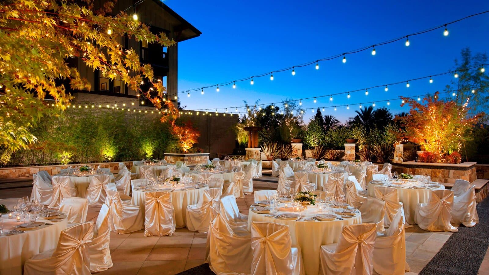 An outdoor terrace at The Westin Verasa Napa set with draped round tables under string lights at dusk in Napa, CA