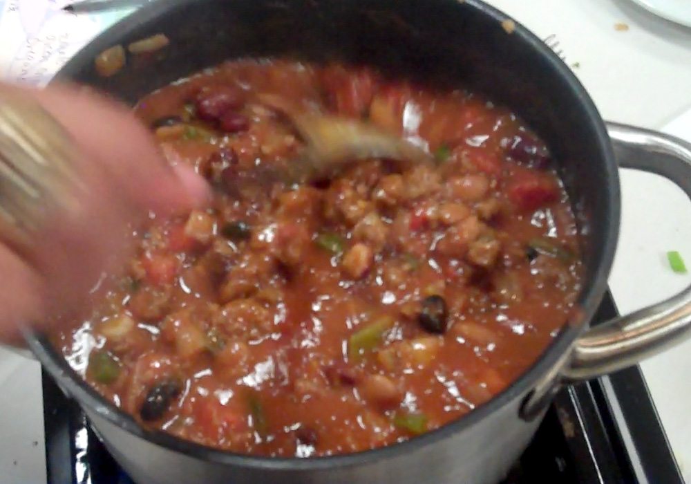 A person stirring chili in a pot on top of a stove.
