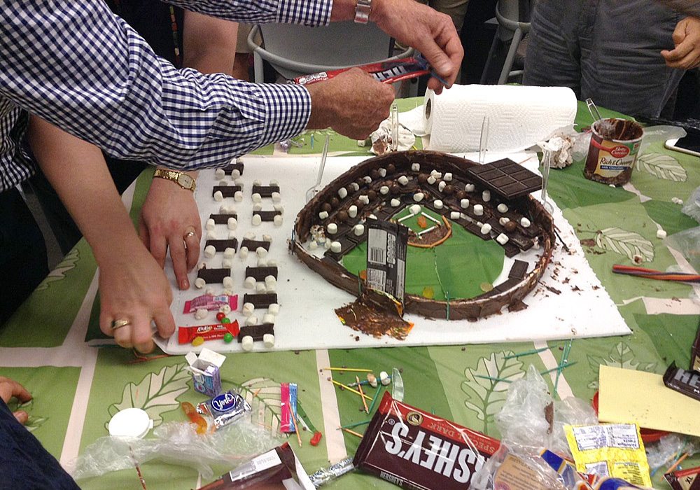 A group of people standing around a cake with a baseball field on it.