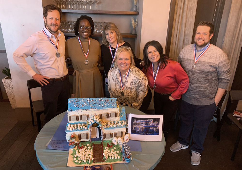 A group of people posing in front of a gingerbread house.