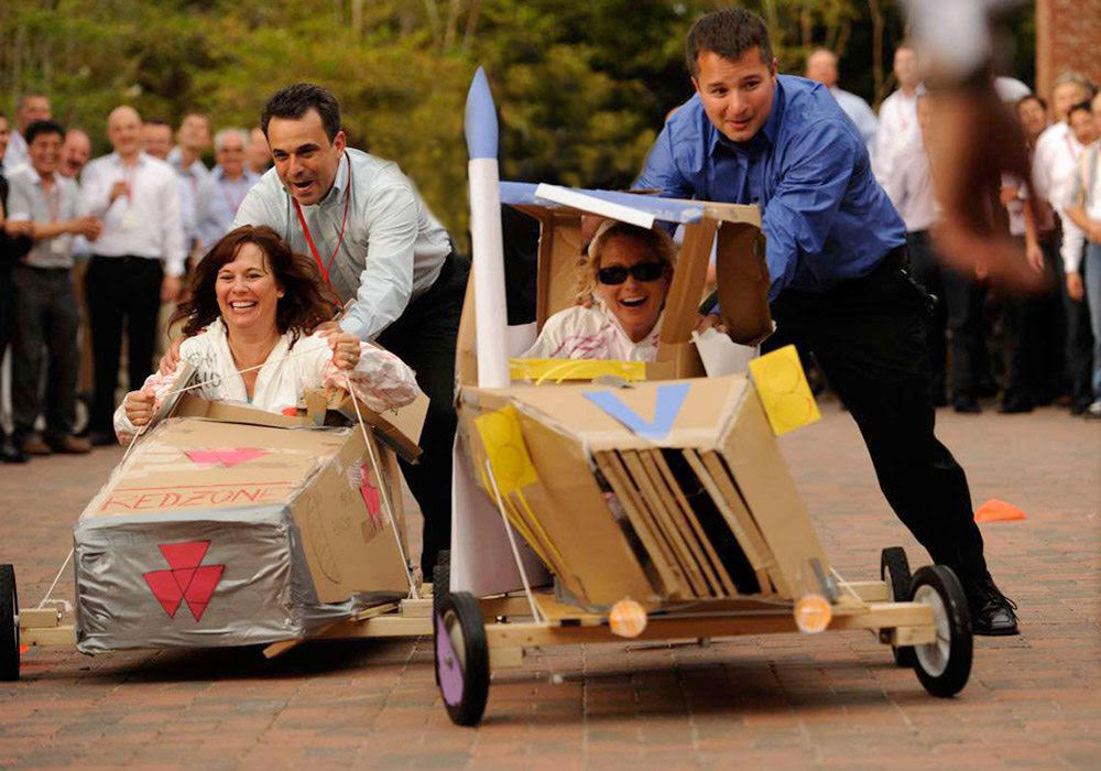 A group of people are racing cardboard cars in a parking lot.