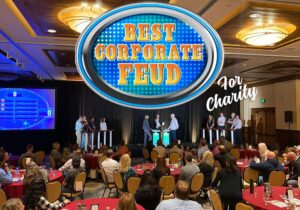 People competing in a corporate-themed game show event for charity, with an audience watching. A sign above reads "Best Corporate Feud.