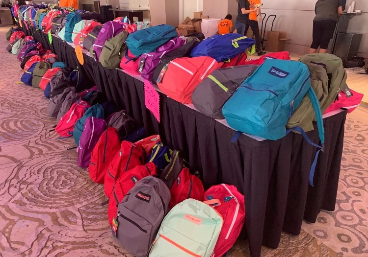 Many backpacks are lined up on a table in a room.
