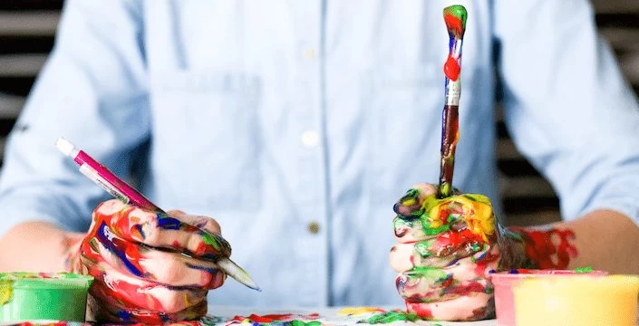 A person holding paint brushes and paints on a table.