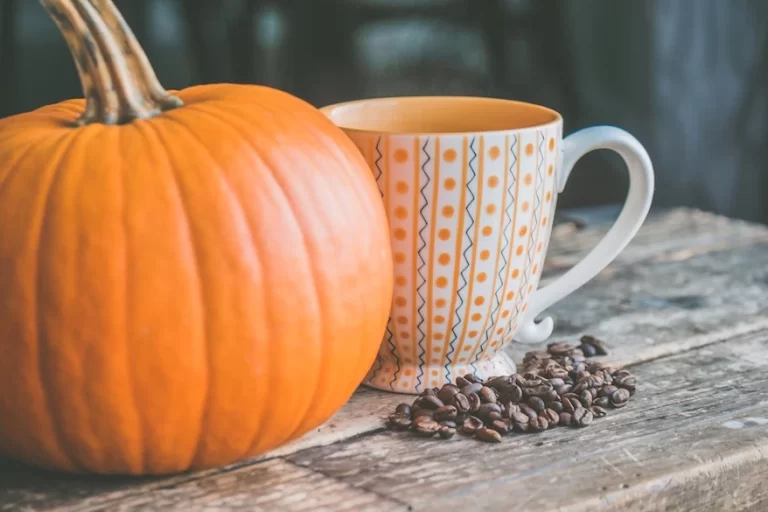 A steaming cup of pumpkin spice coffee sits next to a vibrant orange pumpkin on a rustic wooden table.