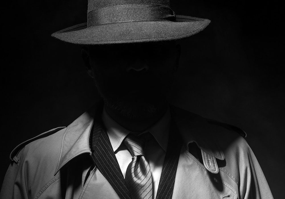 A man in a trench coat and hat is silhouetted against a black background.