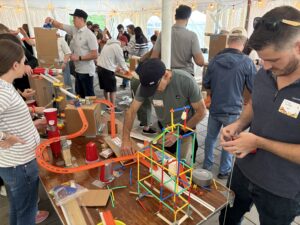 People assemble and test colorful toy structures and tracks on tables at a busy indoor event, surrounded by others working on similar projects.