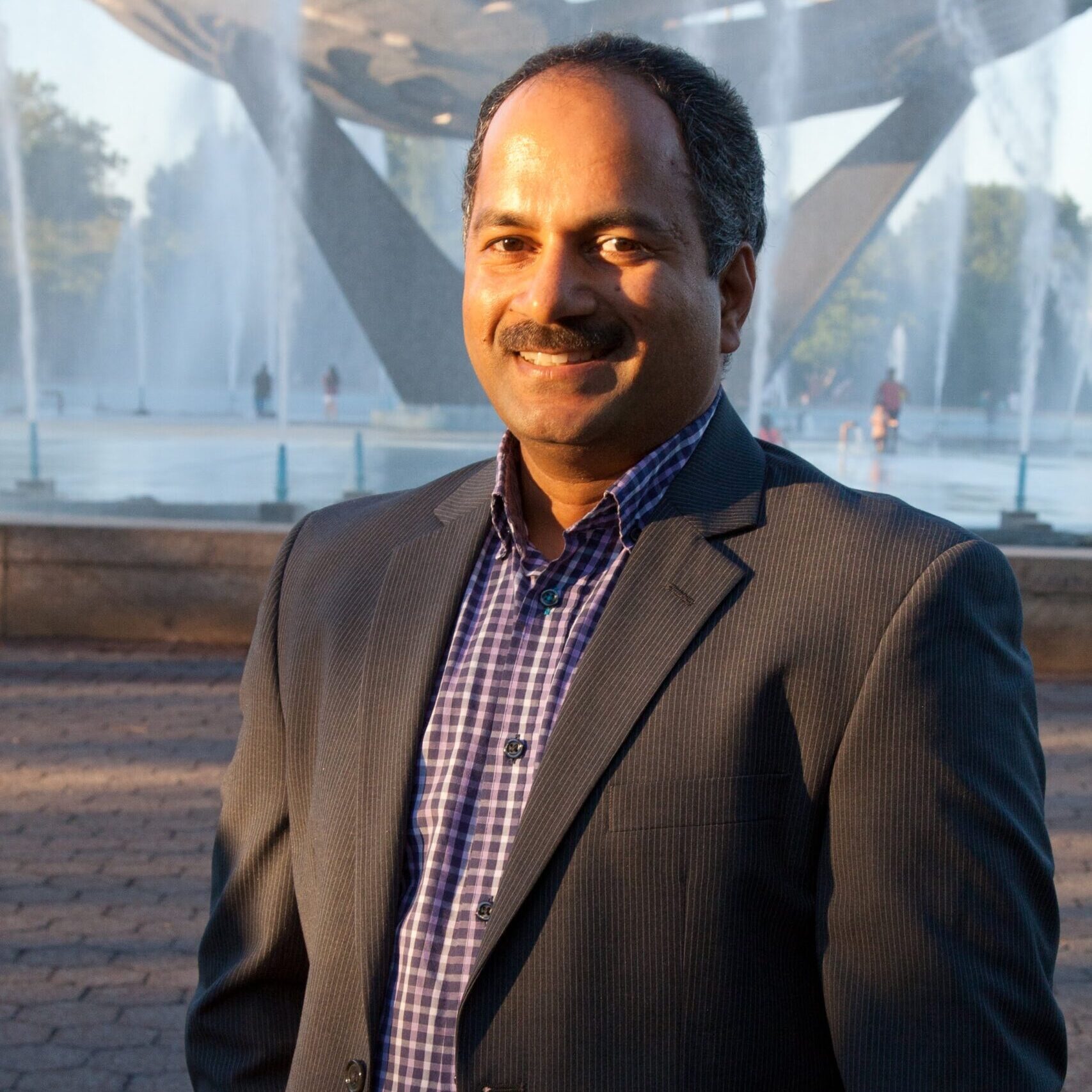 A man in a suit stands outdoors in front of a fountain and a large metal sculpture, smiling at the camera.