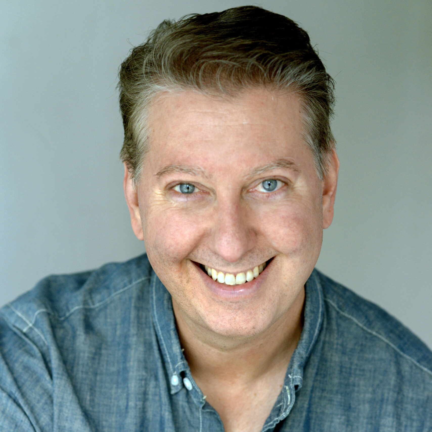 A middle-aged man with light skin and short brown hair, smiling at the camera, wearing a blue button-up shirt against a plain background.
