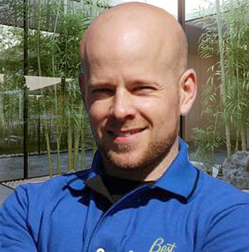 A bald man in a blue shirt standing in front of bamboo.