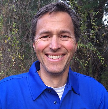 A smiling man in a blue shirt standing in a wooded area.