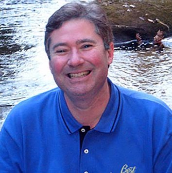 A man in a blue shirt smiling in front of a stream.