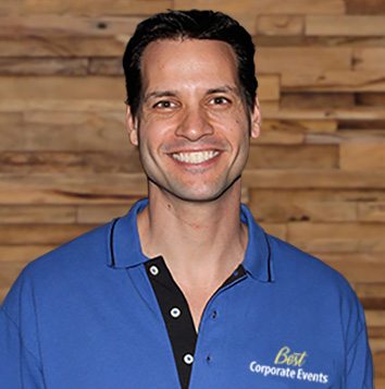 A man in a blue shirt smiling in front of a wooden wall.