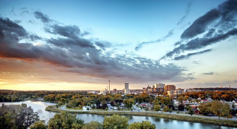 Panoramic view of a cityscape with a river and trees in the foreground, buildings and skyline in the background, and a sky with clouds during sunset.