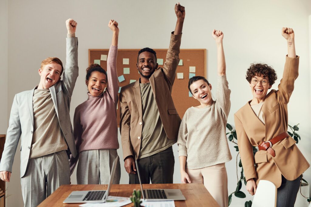 Five people stand in an office, smiling and raising their arms in celebration, embodying the spirit of the best place to work. Two laptops and papers are on the table before them, capturing a moment of achievement and camaraderie.