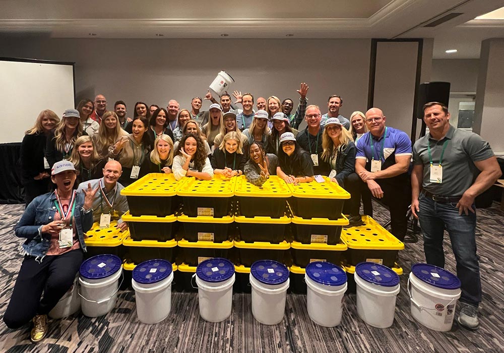 A group of people pose in a room behind stacked yellow boxes and white buckets, some wearing hats, showcasing a hydroponics donation at an indoor event or conference.