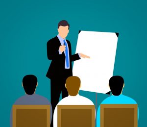 An illustration of a man in a suit giving a presentation to four seated attendees, pointing at a blank flip chart, discussing the latest employee training trends.