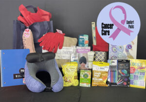 Gift bag with items including a neck pillow, gloves, notebook, snacks, and self-care products. A logo with a pink ribbon and text "Cancer Care Comfort Packs" is visible, offering the perfect blend of support and thoughtfulness in our cancer care packs.