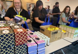 A group of people gathers supplies from a table loaded with various boxed items, placing them into blue cancer care packs.