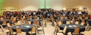 A large group of people is standing together in a spacious conference room with round tables and chairs, facing the camera. The room is well-lit, and the participants appear to be at a formal custom CSR event.