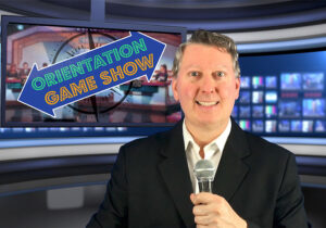 A smiling man in a suit holds a microphone with a television screen behind him displaying the text "Orientation Game Show." The background includes an array of lit monitors.