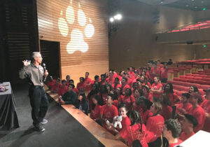 A speaker addresses a large group of people wearing red shirts in an auditorium, with a paw print symbol illuminated on the wall behind him.