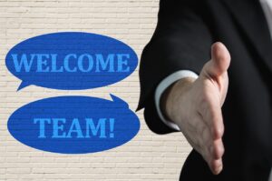 A person in a suit extending their hand for a handshake, with two blue speech bubbles on the left that read "WELCOME TEAM!" against a white brick wall background, setting the stage for a positive employee onboarding experience.