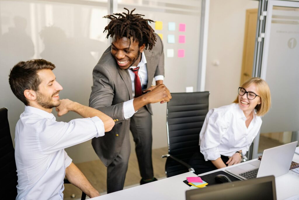 Three colleagues in an office, with one man standing and fist-bumping a seated man, while a woman sitting nearby smiles.