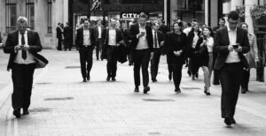 A group of people in business attire walk down a city street, with several individuals looking at their phones.