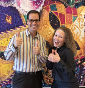 Two people smiling and giving thumbs up in front of a colorful, sequined backdrop.