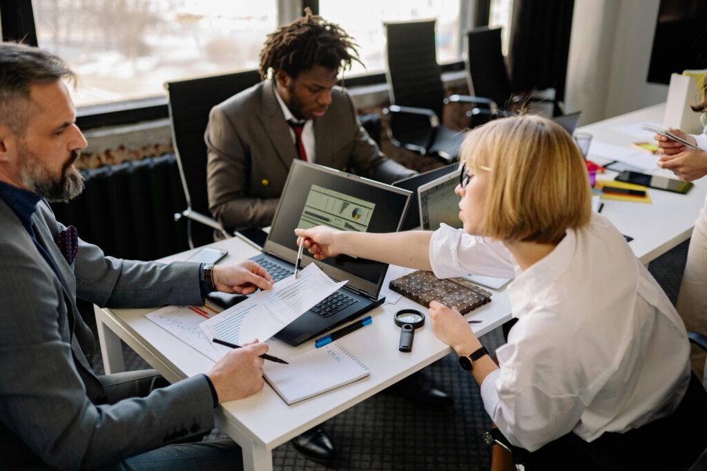 Three colleagues sitting at a desk with laptops and documents, discussing a project. A woman points at a paper with a pen, while two men listen.