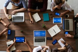 People seated around a wooden table with laptops, tablets, and notebooks are discussing check-in questions for meetings. Devices display blue screens with charts. Coffee cups and a small plant add warmth to the collaborative atmosphere.