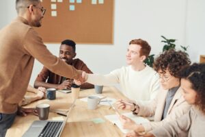 A group of five people sit at a conference table, with two individuals in the center shaking hands in a professional meeting setting.