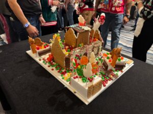 A decorated gingerbread house with candy and cookies sits on a black table, surrounded by people in festive clothing.