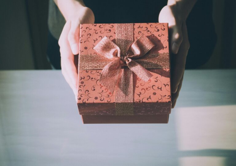 Hands holding a red gift box with a patterned design and a glittery ribbon bow, over a light-colored table.