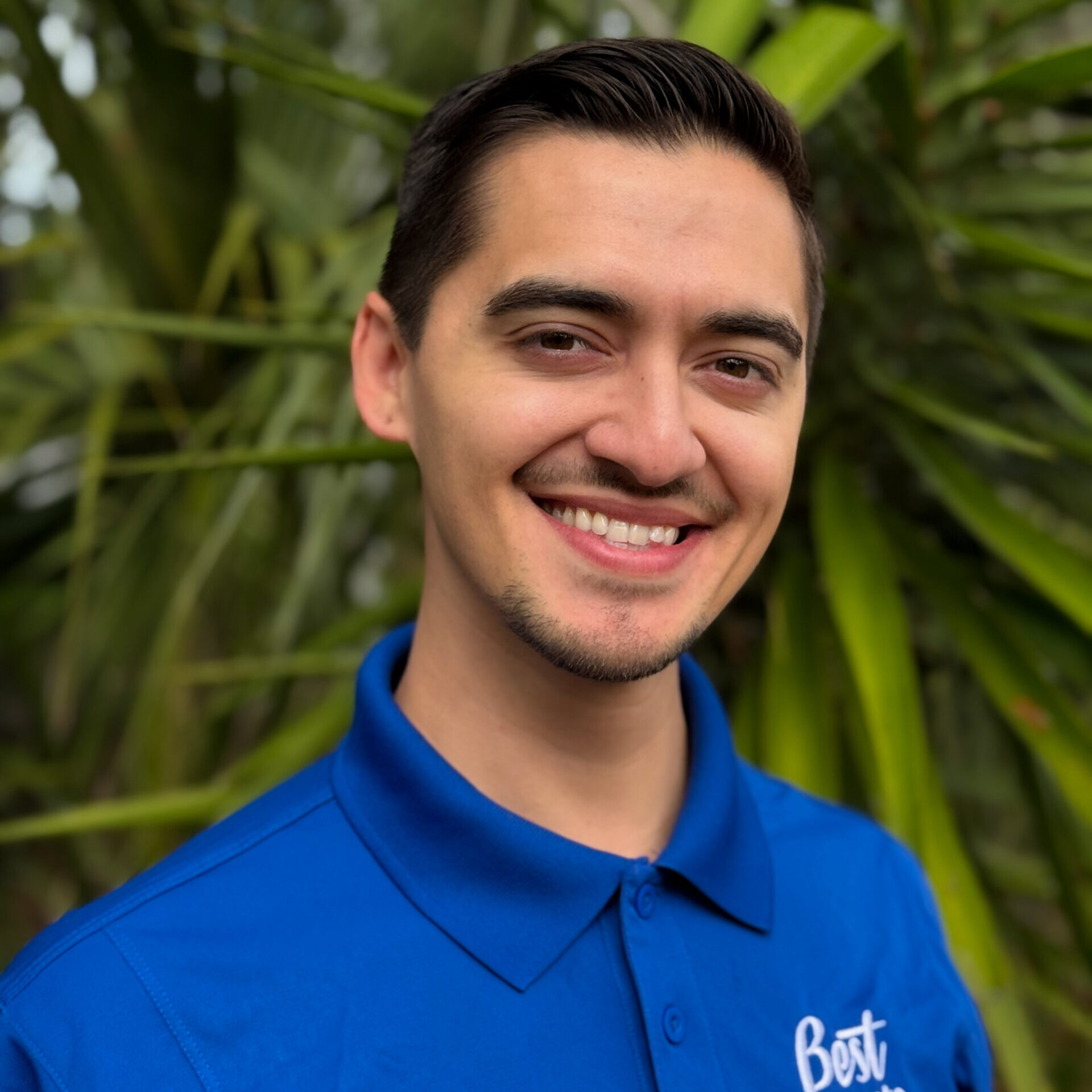 A young man with short dark hair, wearing a blue "Best Corporate Events" polo shirt, smiles while standing in front of green foliage.