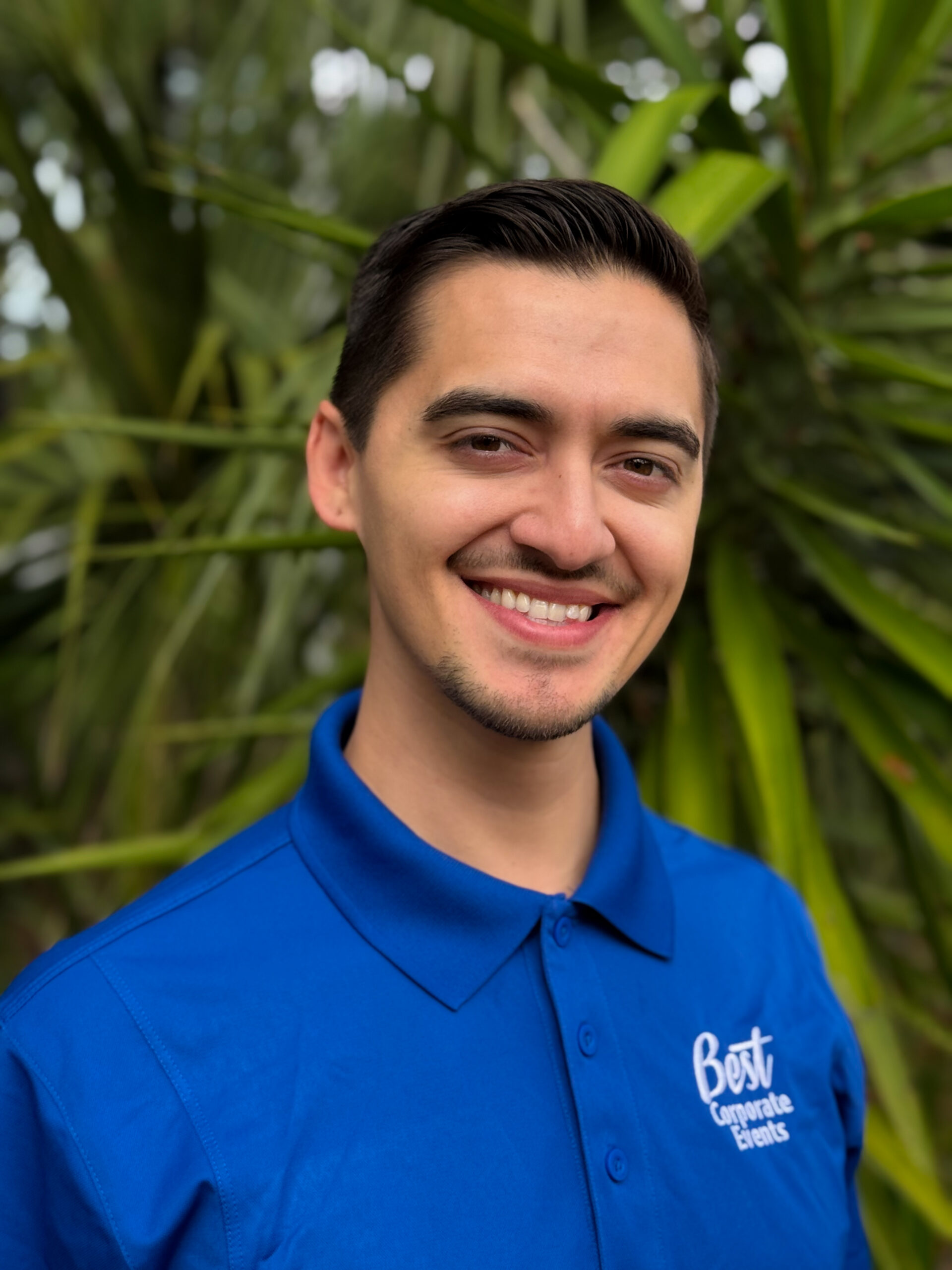 A young man with short dark hair, wearing a blue "Best Corporate Events" polo shirt, smiles while standing in front of green foliage.