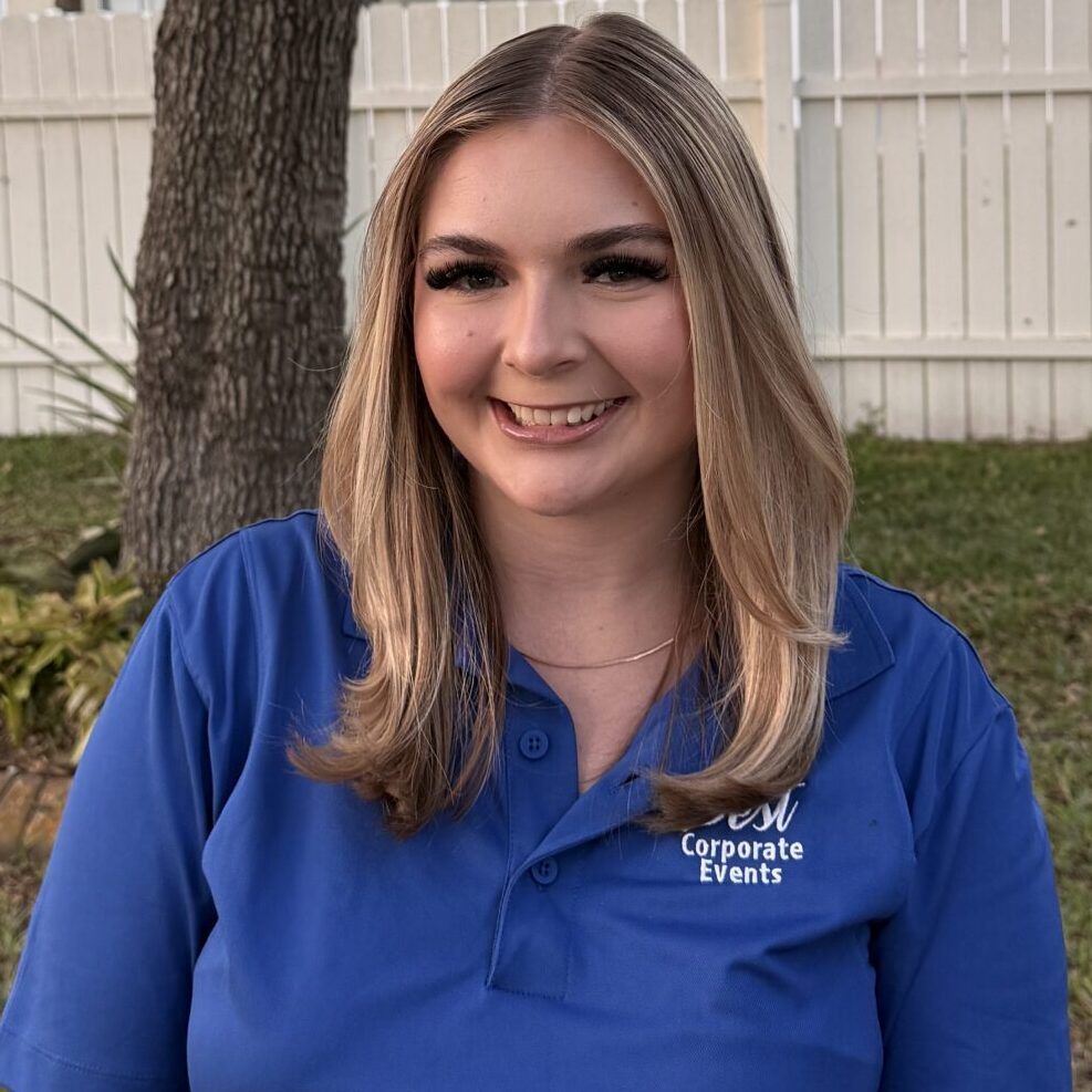 A woman wearing a blue polo shirt stands in a backyard with a tree, birdhouse, white fence, and houses in the background.