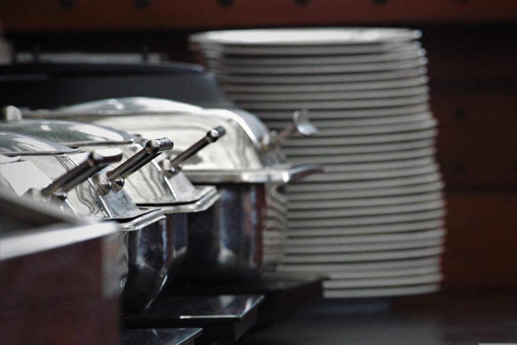 A stack of white plates next to covered stainless steel buffet trays, perfect for serving delicious potluck ideas for work, with a blurred background.