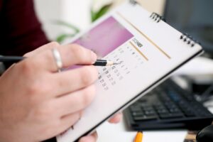 A person holding a pen points to a date on a calendar while sitting at a desk with a keyboard and a computer monitor in the background.