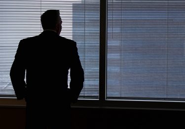 Silhouette of a man in a suit standing in front of large windows with closed blinds, looking outside, reflecting the quiet solitude of workplace loneliness.