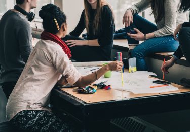 A group of people gathered around a table with papers and markers, engaged in conversation, strategizing how to measure team building success.