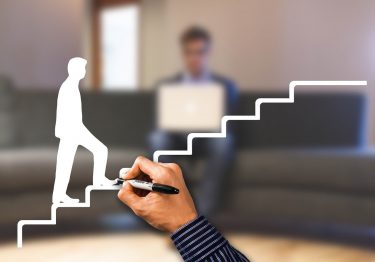 Hand drawing a staircase leading to a man with a laptop, symbolizing progress or career advancement, with a blurred background.