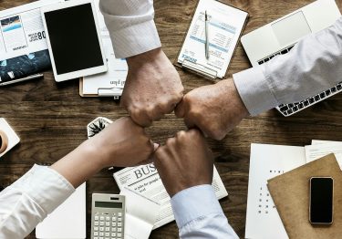 Four individuals fist-bumping over a business meeting table strewn with documents, electronic devices, and coffee cups.